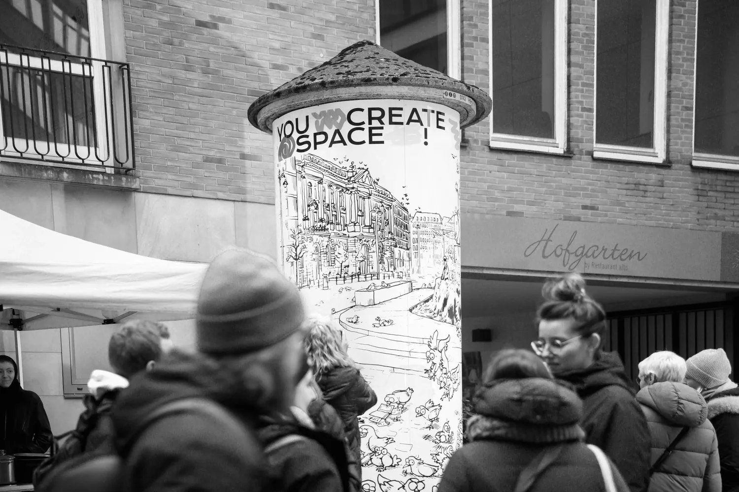 Schwarz-weiß-Fotografie einer Litfaßsäule in der Stadt, beklebt mit einer Illustration voller Tauben. Menschen mit Winterkleidung stehen davor, darüber steht der Schriftzug ‚You create space‘.